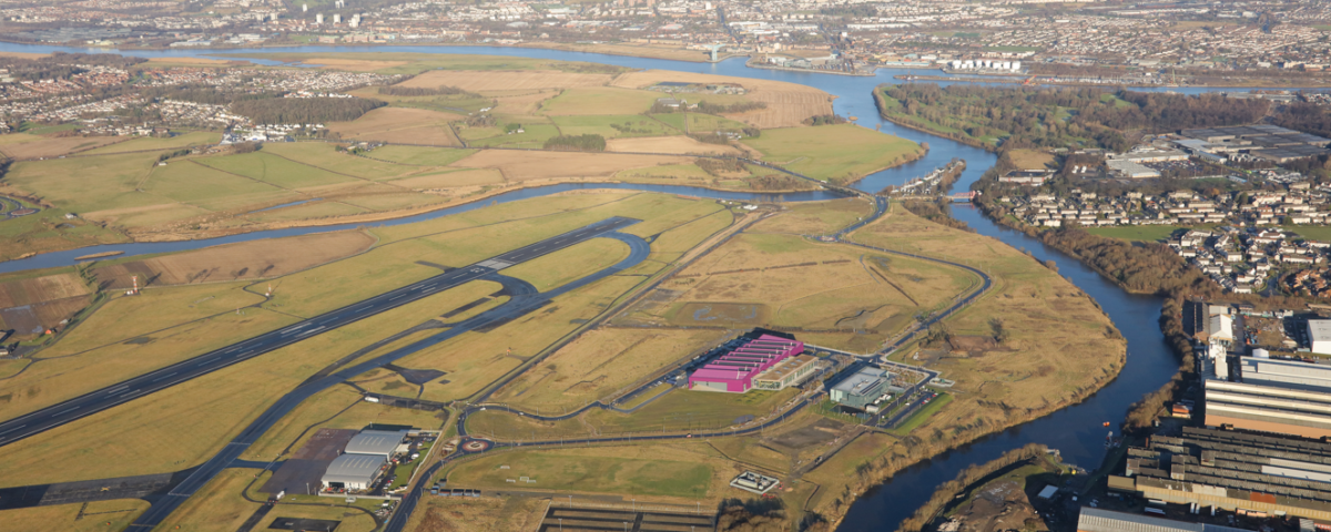 Aerial view of a landscape with a river winding through green fields, industrial buildings, parking lots, and a runway, with a city and hills in the distance under a partly cloudy sky.