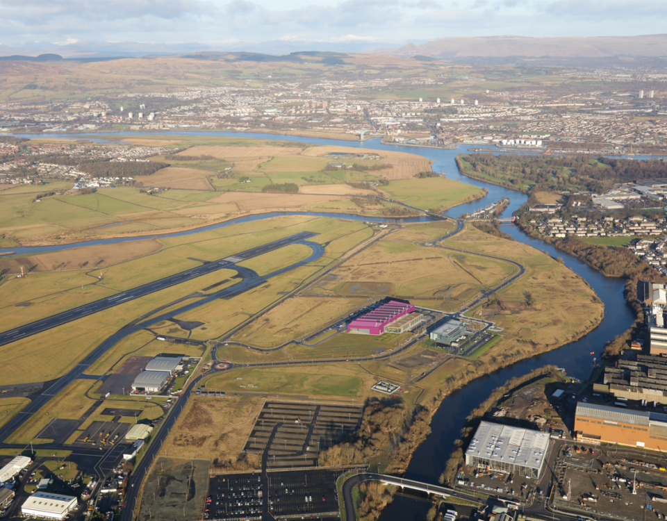 Aerial view of a landscape with a river winding through green fields, industrial buildings, parking lots, and a runway, with a city and hills in the distance under a partly cloudy sky.