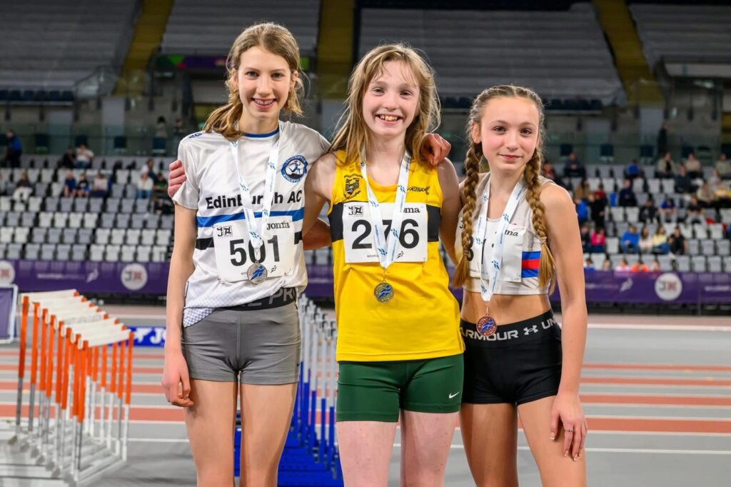 Three young female athletes wearing medals pose together on an indoor track. They stand in front of hurdles, smiling, with an empty stadium and tiered seating in the background.
