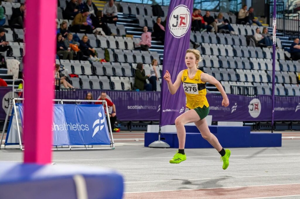 A young athlete in a green and yellow uniform, wearing bib number 276, sprints indoors on a track. Spectators sit in rows of seats, and purple banners with the Scottish Athletics logo are visible in the background.