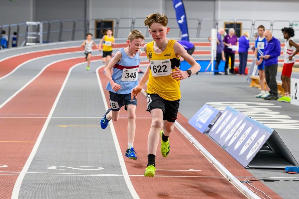 Two boys sprint on an indoor track, with the boy in a yellow jersey (number 622) leading ahead of a boy in a white jersey (number 348). Officials and other athletes stand near the track in the background.