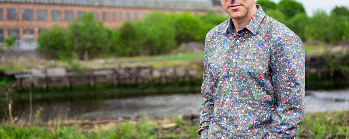 A bald man in a colorful floral shirt stands outdoors near a river, with green bushes and a large brick building visible in the background. He has his hands in his pockets and looks at the camera.