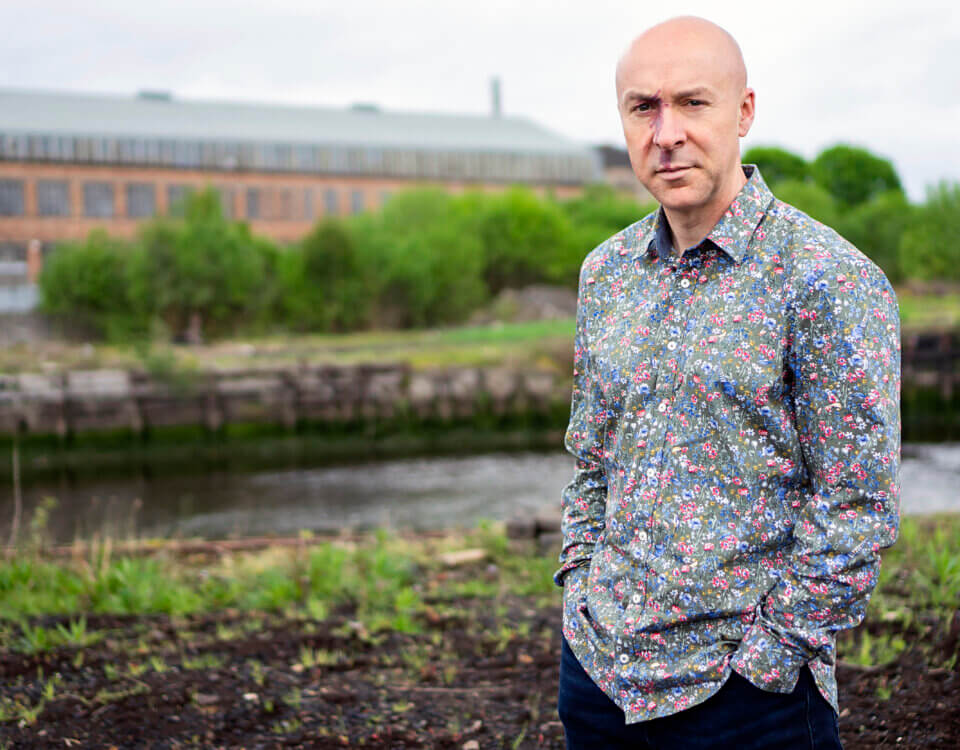 A bald man in a colorful floral shirt stands outdoors near a river, with green bushes and a large brick building visible in the background. He has his hands in his pockets and looks at the camera.