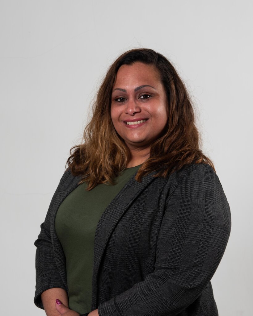 A woman with wavy brown hair, wearing a green top and a gray blazer, smiles while posing in front of a plain white background.