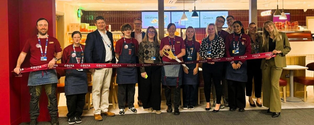 A group of people, including staff in uniforms and others in business attire, stand in front of a Costa Coffee shop holding a long red ribbon, suggesting a ribbon-cutting event or store opening.