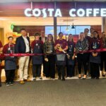A group of people, including staff in uniforms and others in business attire, stand in front of a Costa Coffee shop holding a long red ribbon, suggesting a ribbon-cutting event or store opening.