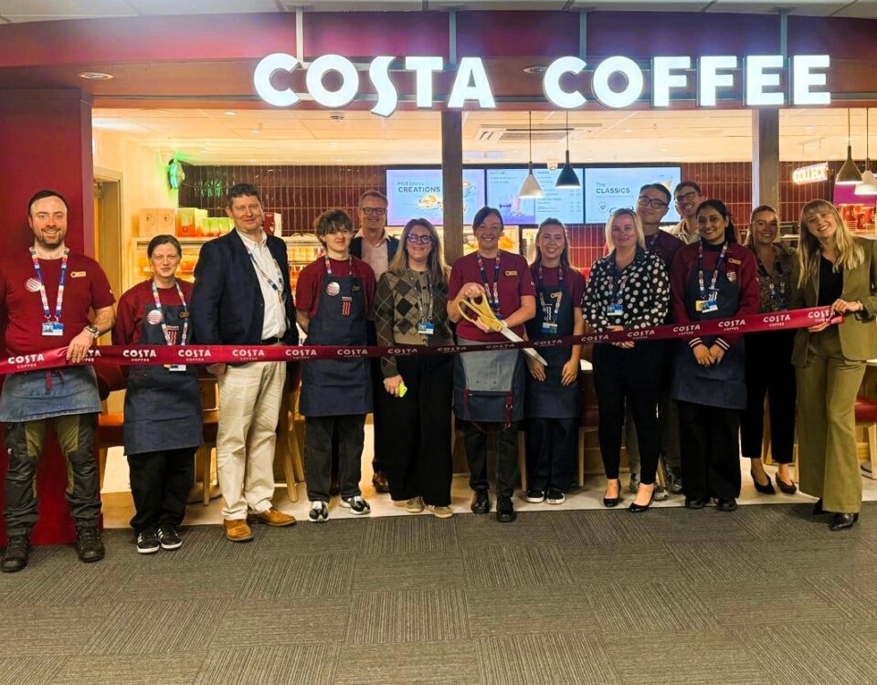 A group of people, including staff in uniforms and others in business attire, stand in front of a Costa Coffee shop holding a long red ribbon, suggesting a ribbon-cutting event or store opening.