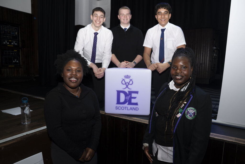 Five students in school uniforms stand around a lit Duke of Edinburgh's Award (DofE) Scotland sign, smiling at the camera in an auditorium setting.