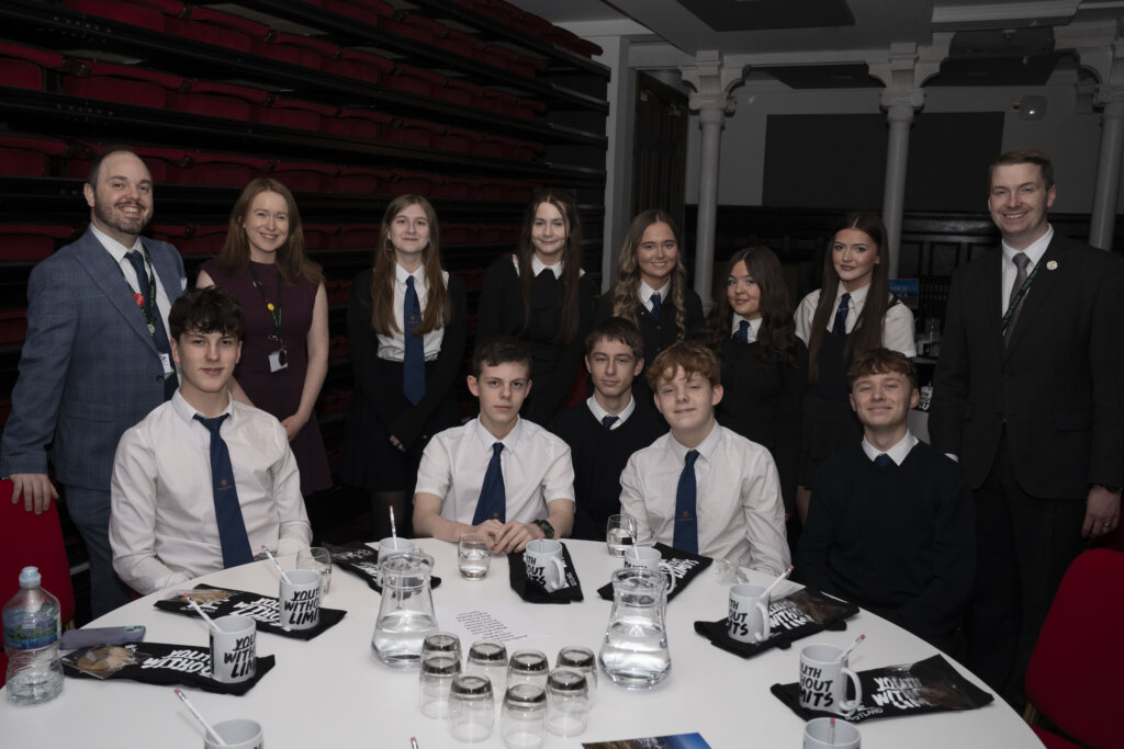 A group of students in school uniforms and four adults in formal wear pose around a round table set with water pitchers, glasses, and booklets in a conference room with red tiered seating in the background.