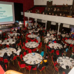 A large event hall with round tables filled with people watching a presentation. A screen displays "David Knox, Duke of Edinburgh Co-ordinator, Mary Russell School." Some people are seated on a balcony above.