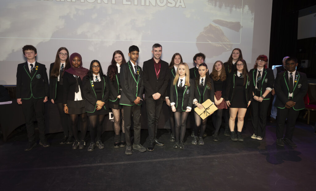 A group of students in black blazers with green trim stand in two rows on a stage with an adult, posing for a photo in front of a screen, some holding folders or certificates.