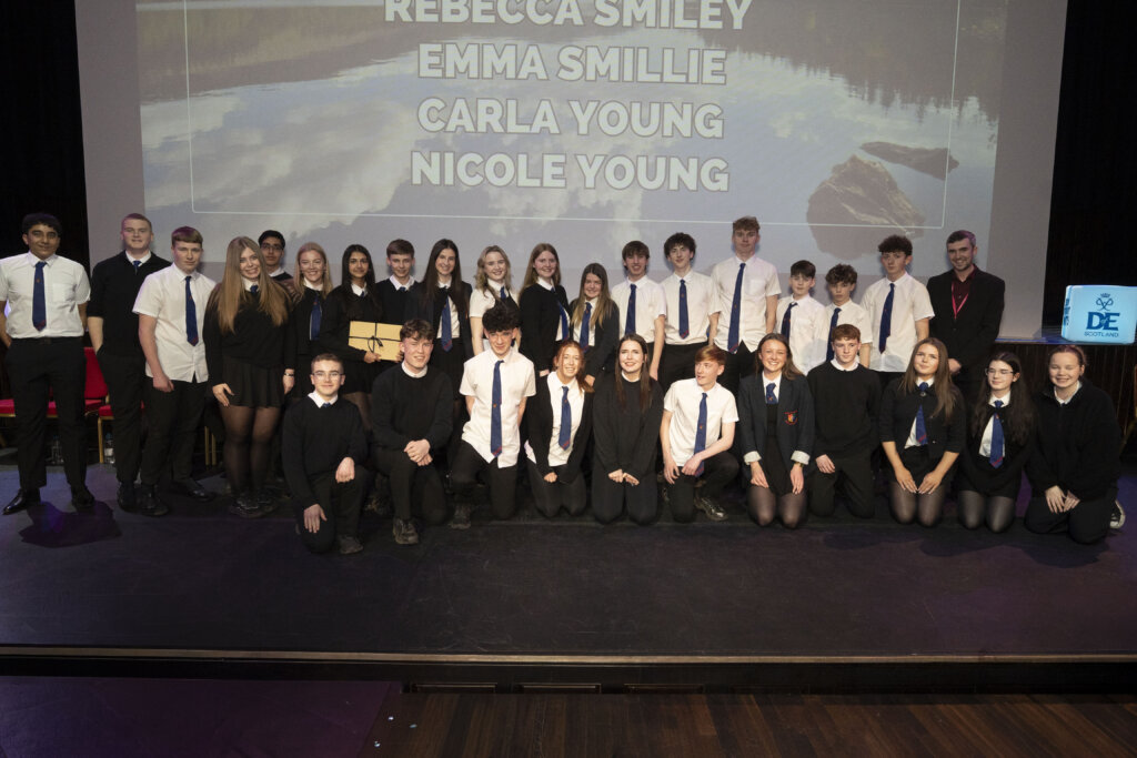 A large group of students and two adults pose on a stage, dressed in school uniforms. Behind them is a screen displaying names: Rebecca Smiley, Emma Smillie, Carla Young, and Nicole Young.