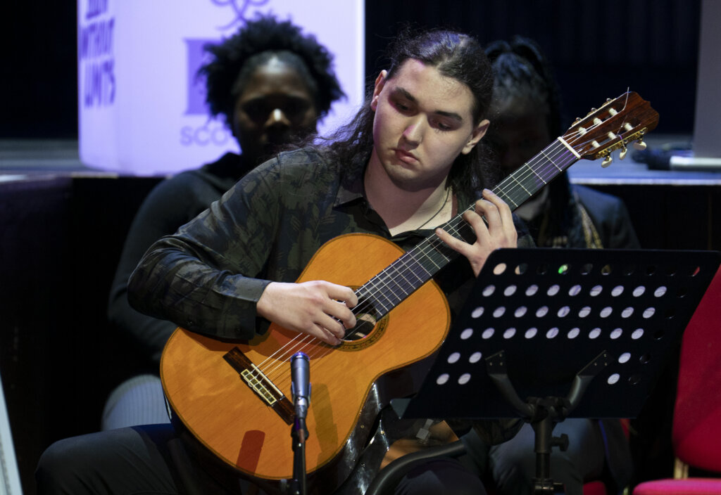 A person with long dark hair plays a classical guitar on stage, focused on the performance. A music stand with sheet music and a microphone are in front of them, with two more people seated in the background.