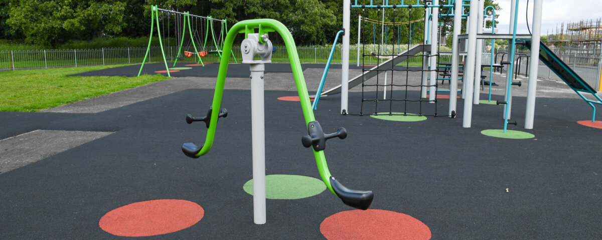 A playground features swings, climbing structures, and a green seesaw-style apparatus on a black rubber surface with red and green circular markings, surrounded by grass and trees.