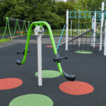 A playground features swings, climbing structures, and a green seesaw-style apparatus on a black rubber surface with red and green circular markings, surrounded by grass and trees.