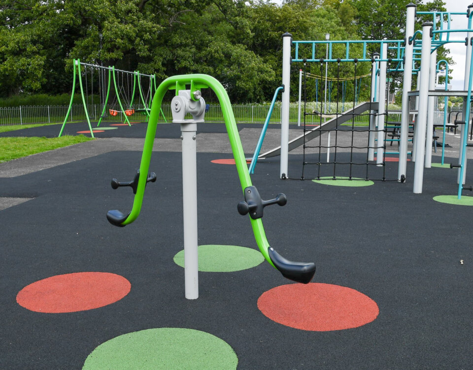 A playground features swings, climbing structures, and a green seesaw-style apparatus on a black rubber surface with red and green circular markings, surrounded by grass and trees.