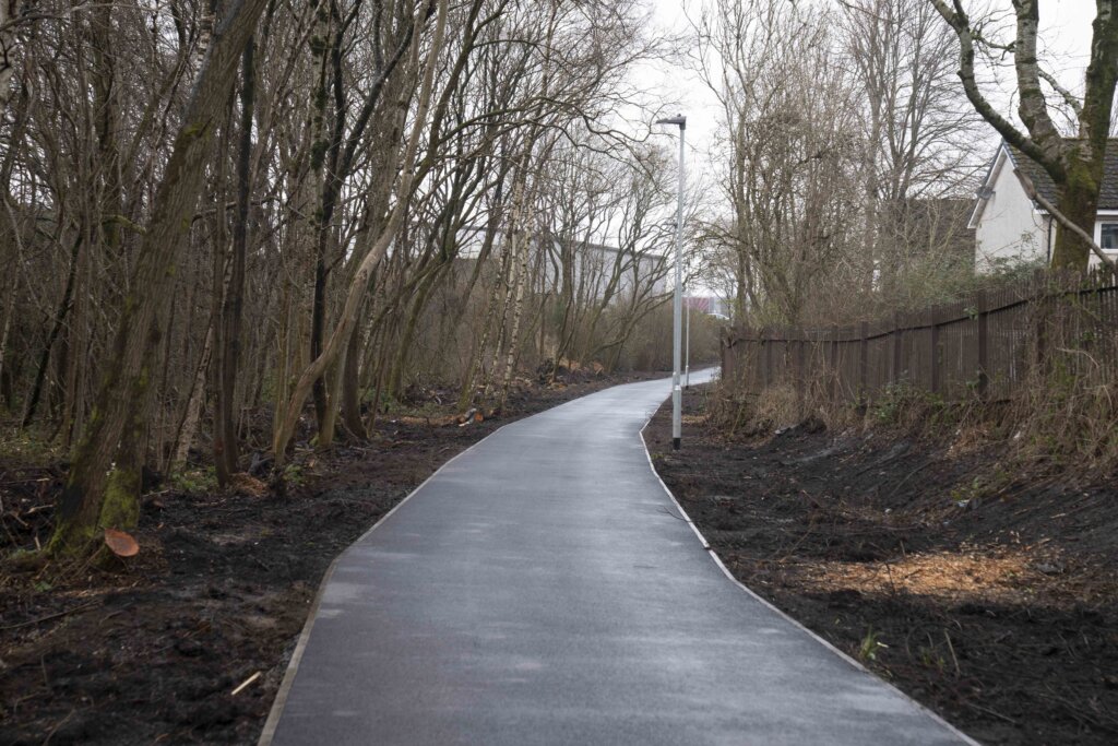 A newly paved path curves through a leafless, wooded area, bordered by a fence and houses on the right, with lamp posts lining the path under a cloudy sky.