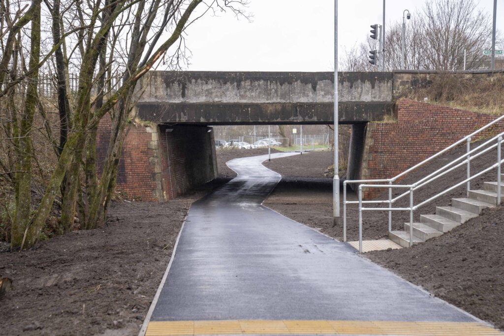 A newly paved path curves under a short brick and concrete bridge on a cloudy day, with bare trees on the left and metal railings with steps on the right. The surroundings are mostly bare soil.