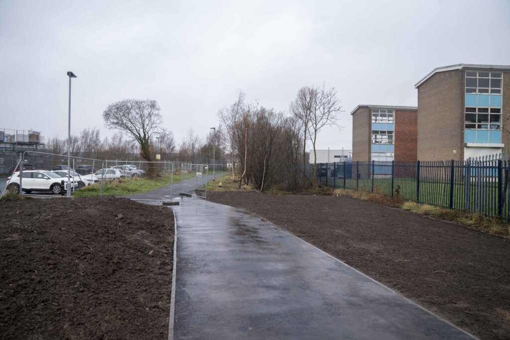 A wet, paved pathway runs through a muddy, fenced area near parked cars and two modern, brick buildings on a cloudy day. Sparse leafless trees line the path, and the sky is overcast.