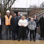 A group of adults and schoolchildren stand outside on a newly paved path near an underpass, some smiling at the camera. One man wears a safety vest and helmet; others are dressed in business or school attire.