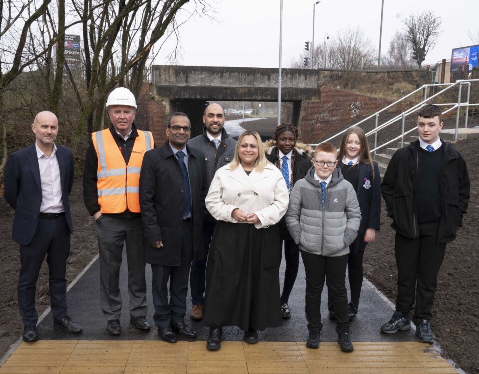 A group of adults and schoolchildren stand outside on a newly paved path near an underpass, some smiling at the camera. One man wears a safety vest and helmet; others are dressed in business or school attire.