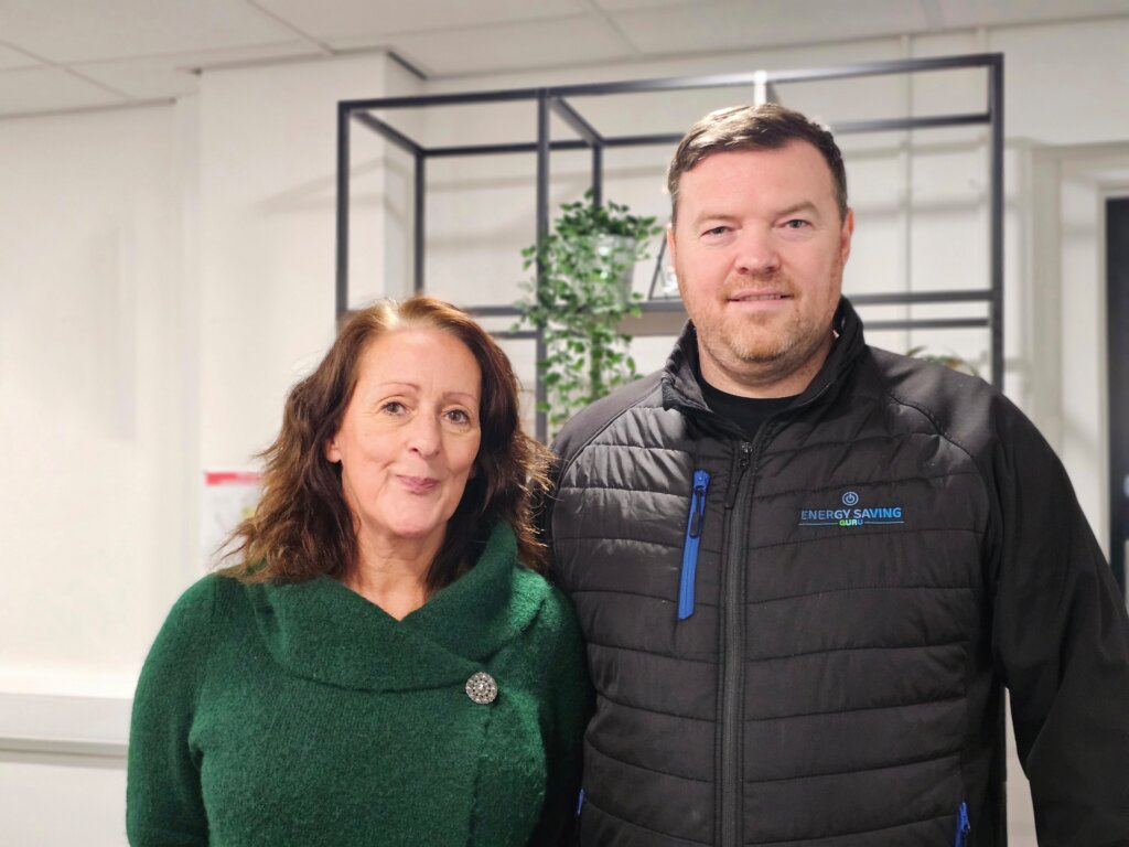 Two adults standing indoors, a woman in a green sweater with a decorative pin and a man in a black "Energy Saving" jacket. A metal shelf with plants and a white wall are in the background.