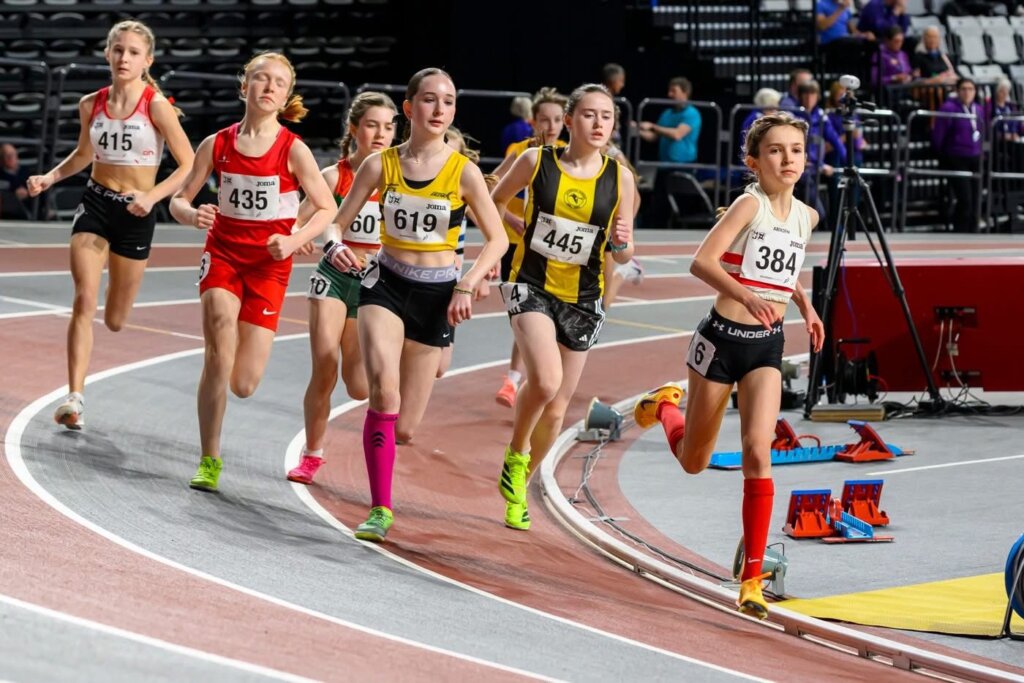 A group of young female athletes run on an indoor track during a race, with one runner in the lead and others closely following behind. Spectators and equipment are visible in the background.