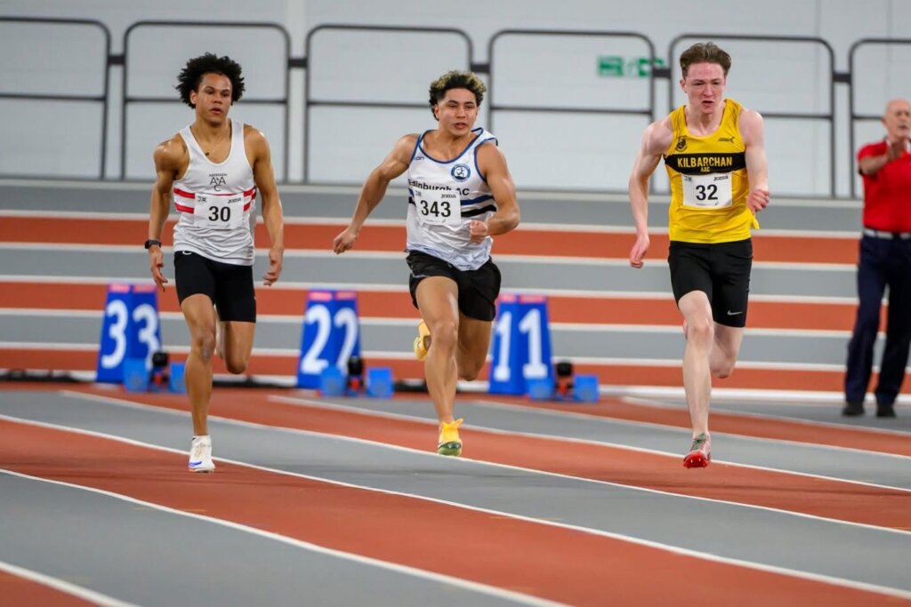 Three male athletes sprint on an indoor track, each in different uniforms. The runner in lane 4 (yellow) leads, while the others (in white jerseys) follow closely behind. A person in red stands in the background.