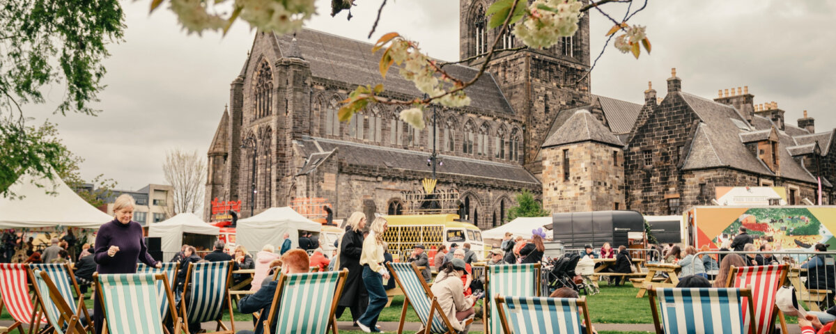 People relaxing on striped deck chairs at an outdoor event with food stalls, in front of a large historic stone church. Flowering branches frame the scene, and the sky is overcast.