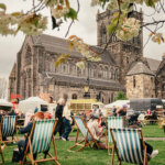 People relaxing on striped deck chairs at an outdoor event with food stalls, in front of a large historic stone church. Flowering branches frame the scene, and the sky is overcast.