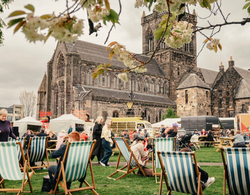People relaxing on striped deck chairs at an outdoor event with food stalls, in front of a large historic stone church. Flowering branches frame the scene, and the sky is overcast.