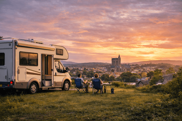 A campervan is parked on a grassy hill overlooking a town at sunset. Two people sit in camping chairs by a table, enjoying the scenic view with a large church or cathedral visible in the distance.