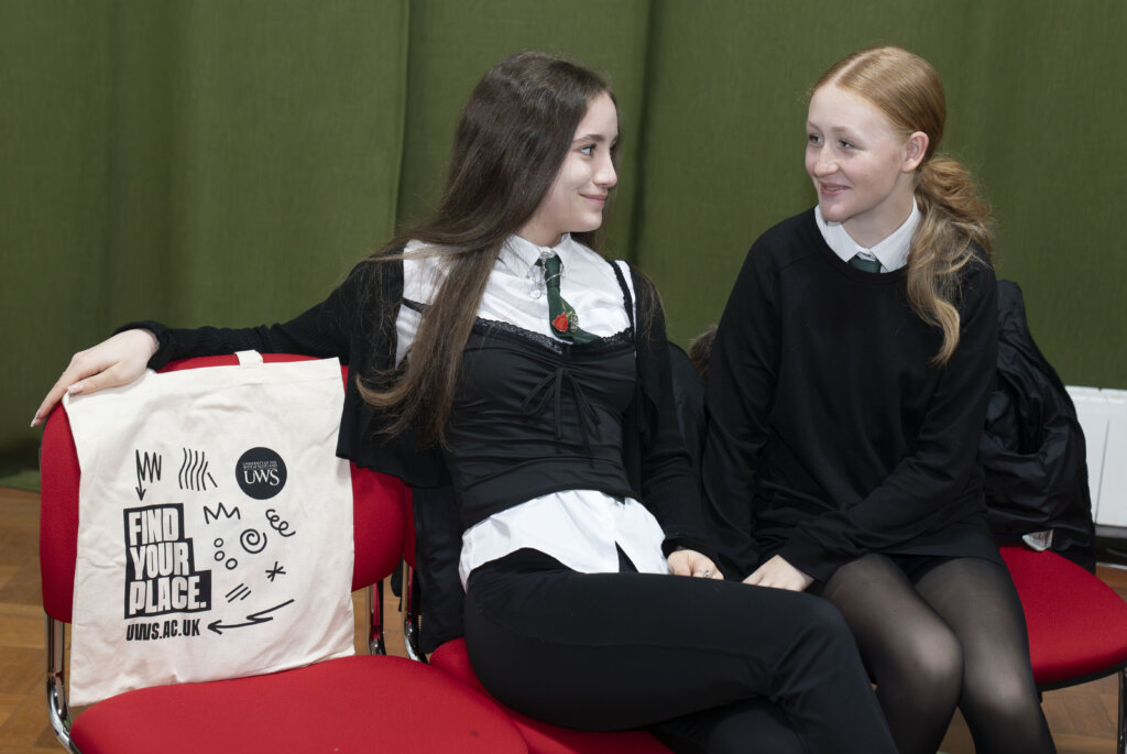 Two young women in school uniforms sit on red chairs, smiling at each other. A beige tote bag with the text "FIND YOUR PLACE UWS AC UK" is on the chair beside them. A green curtain is in the background.