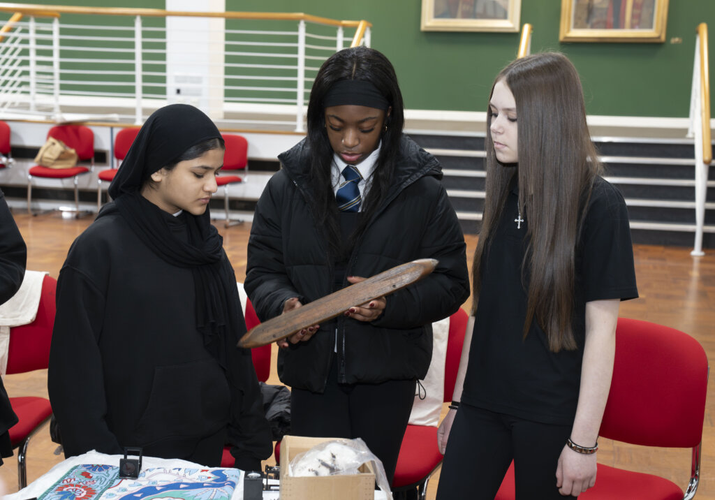 Three teenage girls stand together indoors. One girl in the center holds and examines a wooden artifact while the other two watch. They are surrounded by red chairs and various items on a table.