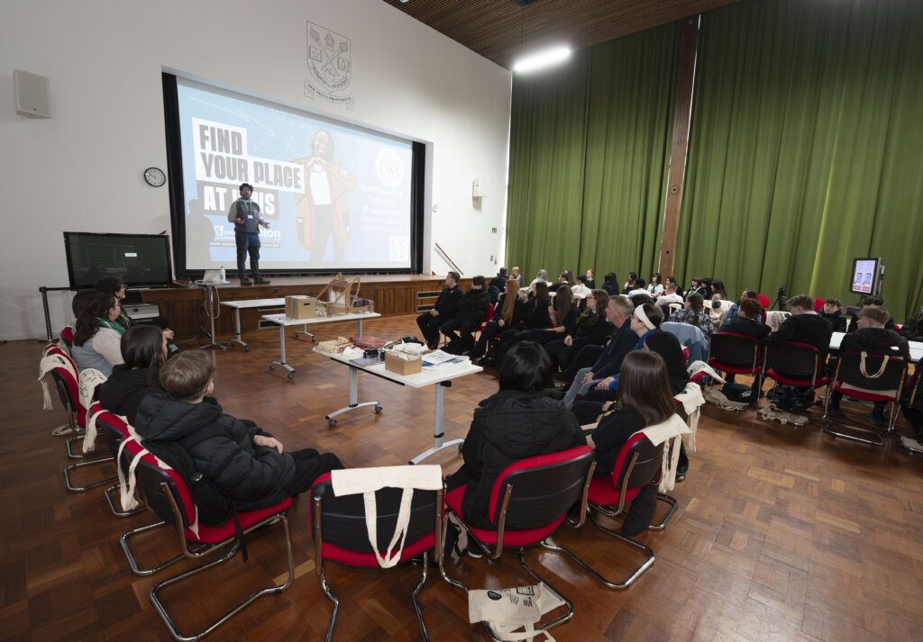 A speaker stands on stage addressing an audience seated in rows in a large hall. A presentation slide behind reads “Find Your Place at UHS.” Tables and bags are placed near the front. Green curtains cover one wall.