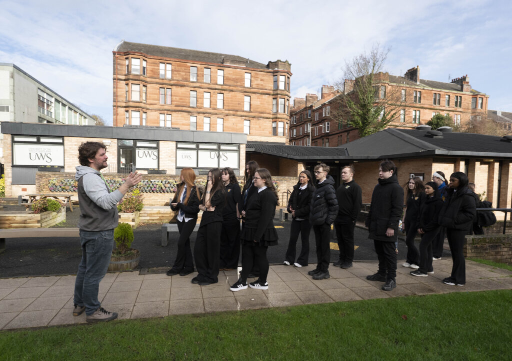 A man stands outside speaking to a group of students dressed in black uniforms, gathered in front of a building with "UWS" signs and red brick buildings in the background.