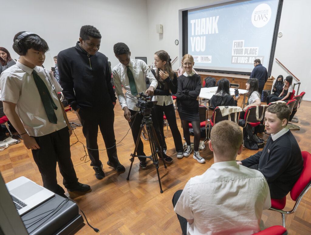 A group of students, some in uniform, gather around a video camera in a classroom while others sit at tables. A large screen in the background reads "THANK YOU! FIND YOUR PLACE AT UWS.SCOT.