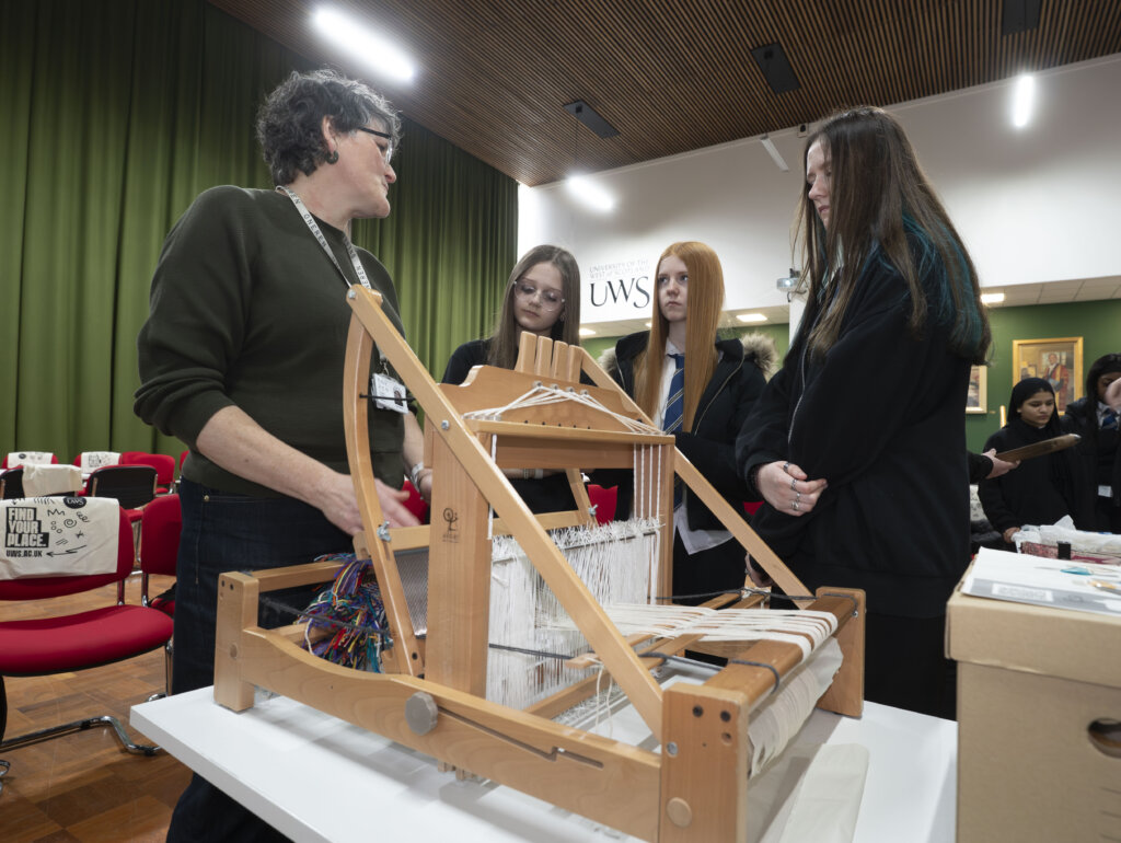 A person demonstrates weaving on a loom to three attentive students in school uniforms. They are in a brightly lit room with green curtains, red chairs, and a University of the West of Scotland (UWS) sign in the background.
