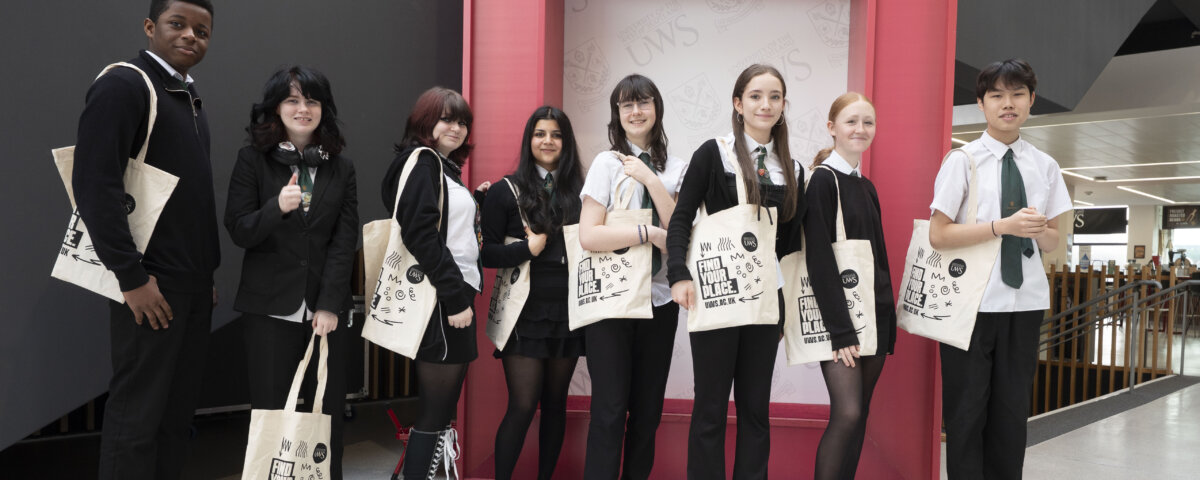 A group of eight students in school uniforms stand smiling with tote bags in front of a red University of the West of Scotland frame inside a modern building.