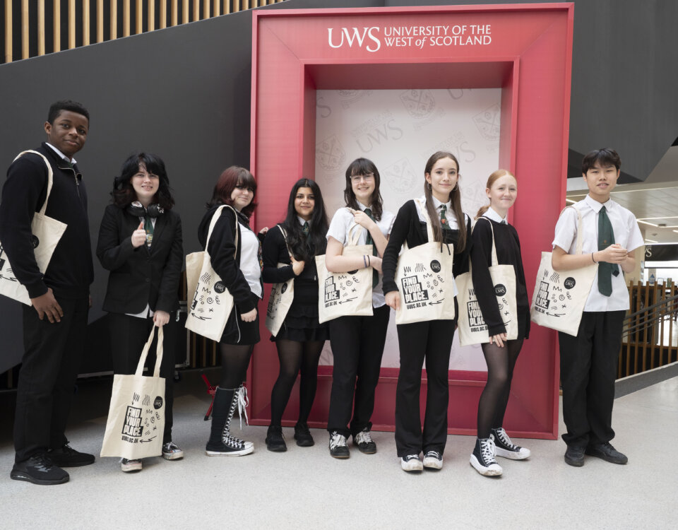 A group of eight students in school uniforms stand smiling with tote bags in front of a red University of the West of Scotland frame inside a modern building.