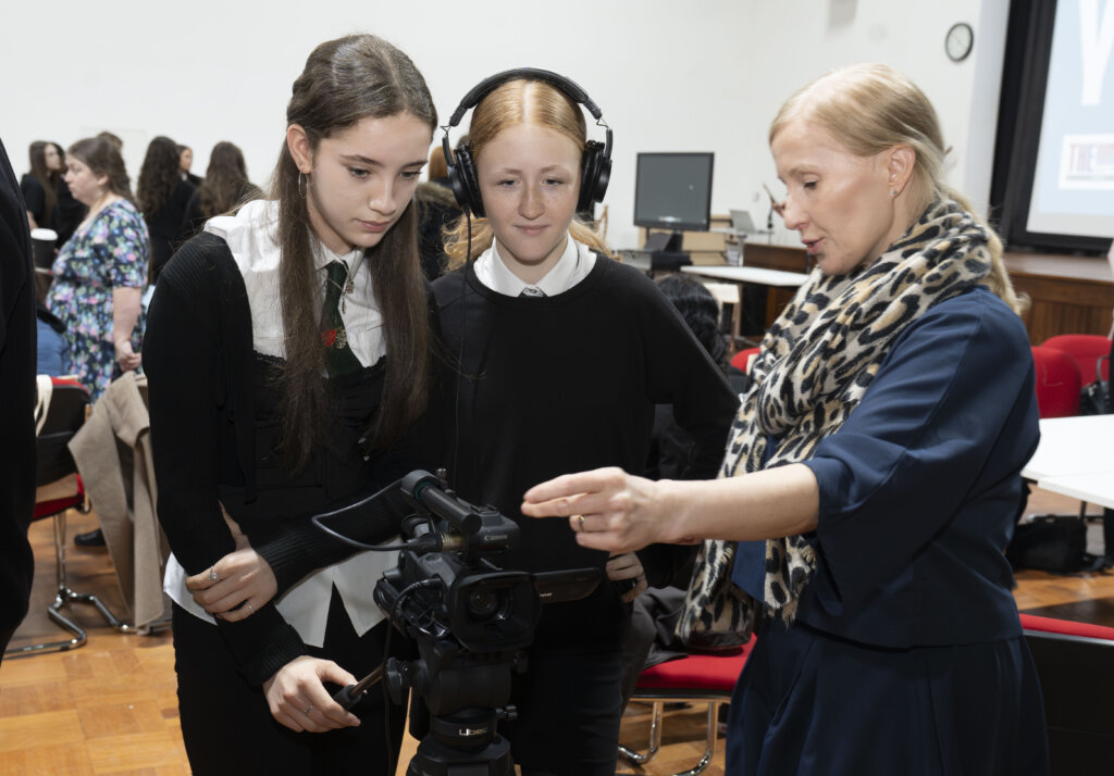 A woman instructs two teenage girls, one wearing headphones, on how to use a video camera in a classroom or workshop setting with other people in the background.