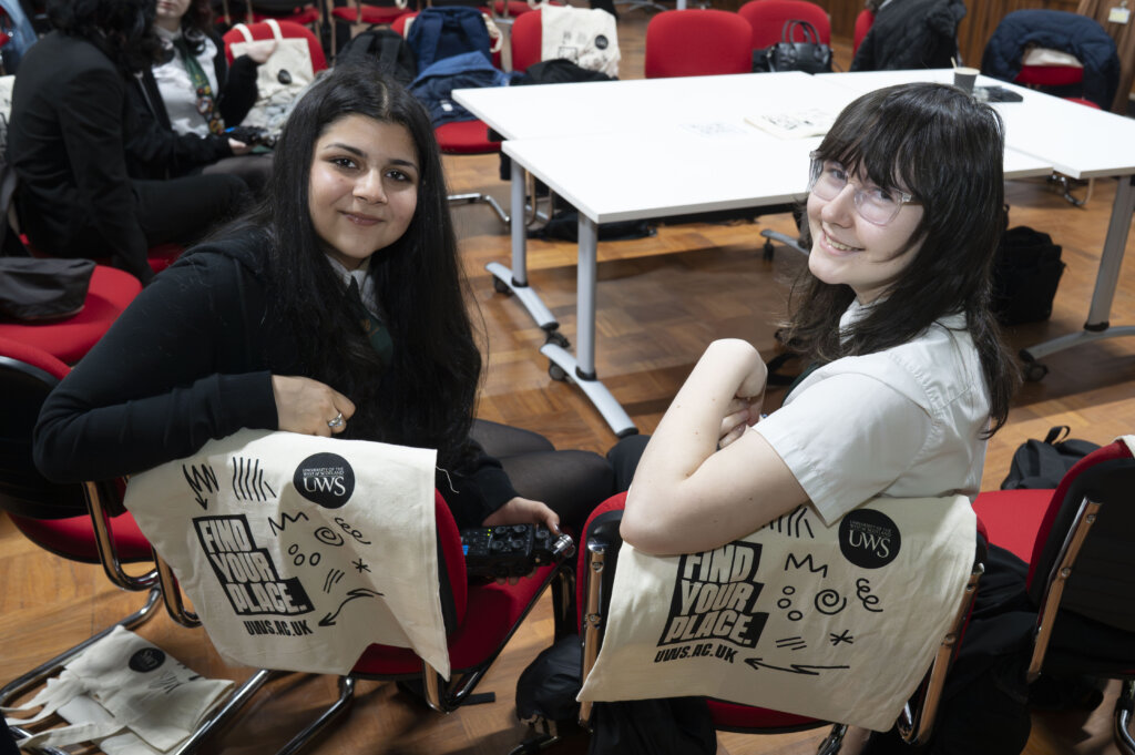 Two young women sit on red chairs, smiling at the camera. Tote bags reading "Find Your Place" are draped over their chairs. They are in a bright room with tables and other people in the background.
