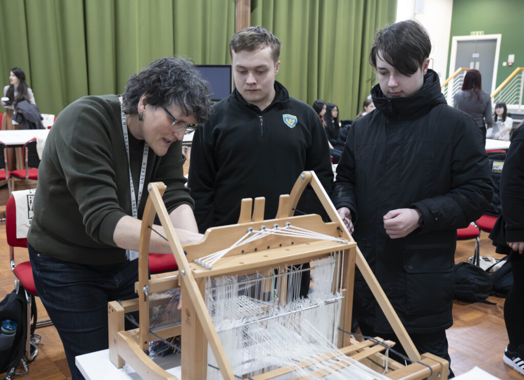 A person demonstrates how to use a wooden weaving loom to two students in a classroom with a green curtain and people in the background.