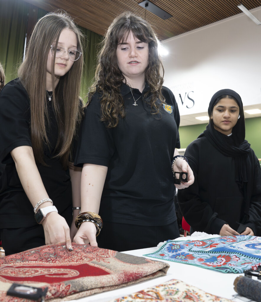 Three young women stand at a table looking at colorful, patterned fabrics. Two of them touch a red and gold textile, while the third, wearing a black headscarf, observes. They appear engaged and interested.