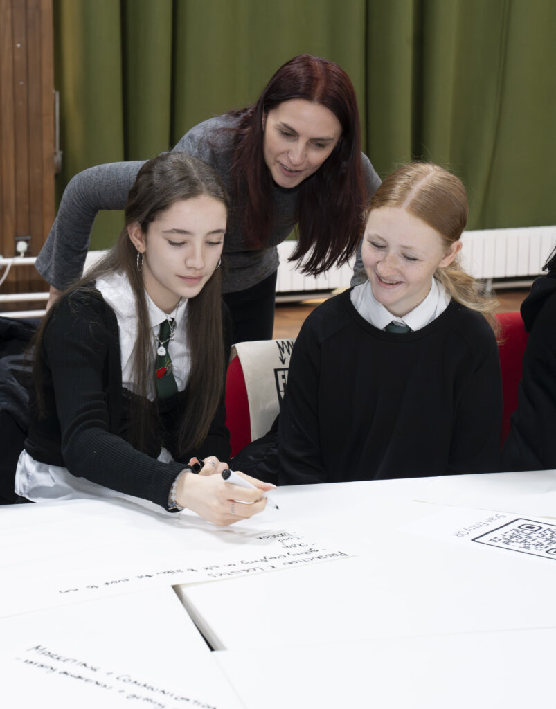 A teacher leans over two students in school uniforms as one student writes on a large sheet of paper, while the other watches and smiles. They appear to be working collaboratively in a classroom setting.