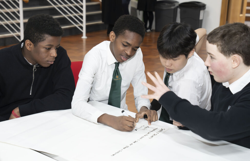 Four boys sit around a table working together, with one writing on a large sheet of paper while another gestures animatedly. They appear focused and engaged in a group activity or discussion.