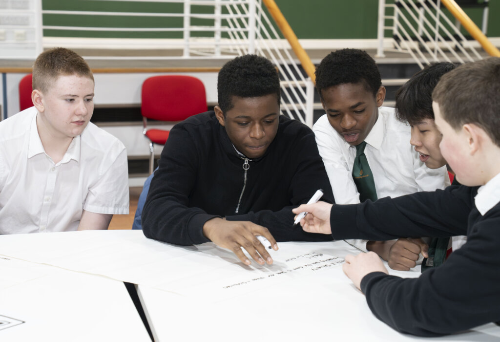 A group of five boys sit around a table in a classroom, collaboratively writing on large sheets of paper. They appear focused and engaged in their group work or discussion.