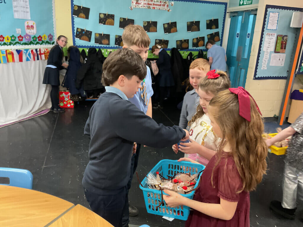 A group of children stand in a classroom, gathered around a blue basket of utensils, with coats hung on hooks and artwork displayed on the walls in the background.
