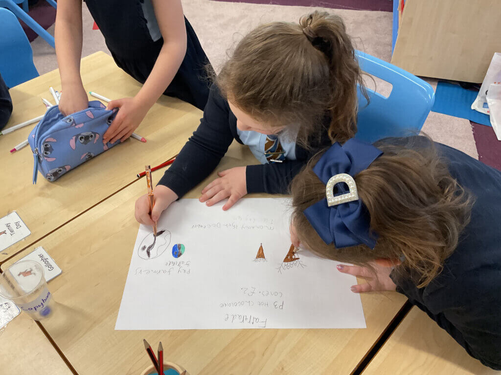 Two young children lean over a table, drawing and writing on a large sheet of paper with colored pencils. One child wears a blue hair bow and both appear focused on their collaborative project.