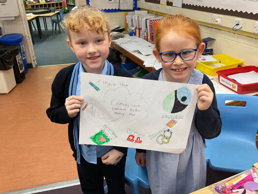 Two young girls in school uniforms smile and hold up a handmade poster about a project. The poster includes colorful drawings and words like “Chocolate,” “Monster,” and “Dinosaurs.” Classroom supplies are visible in the background.